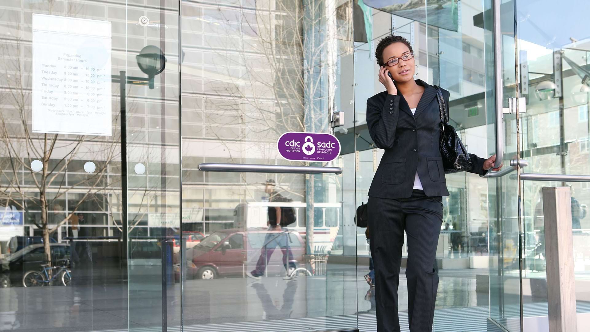 A woman in a black suit talks on her phone while exiting a modern glass building.